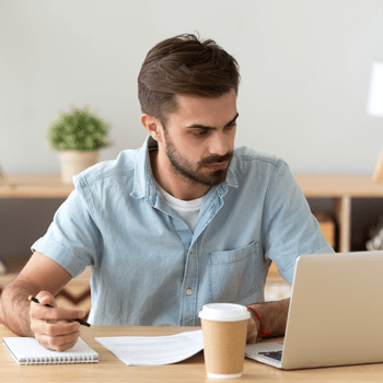 A man sits at a table in front of a laptop. He is holding a pen in his right hand over a spiral notebook. There is a cup of take away coffee on the table.