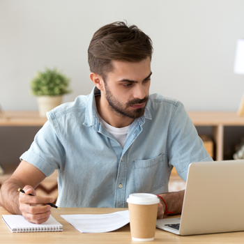 A young man is sitting in front of a laptop taking notes.